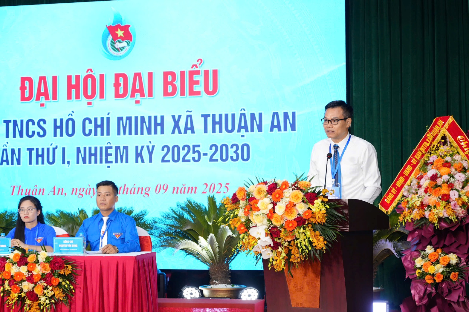 A person standing at a podium with a microphone and a person in a blue shirt  AI-generated content may be incorrect.