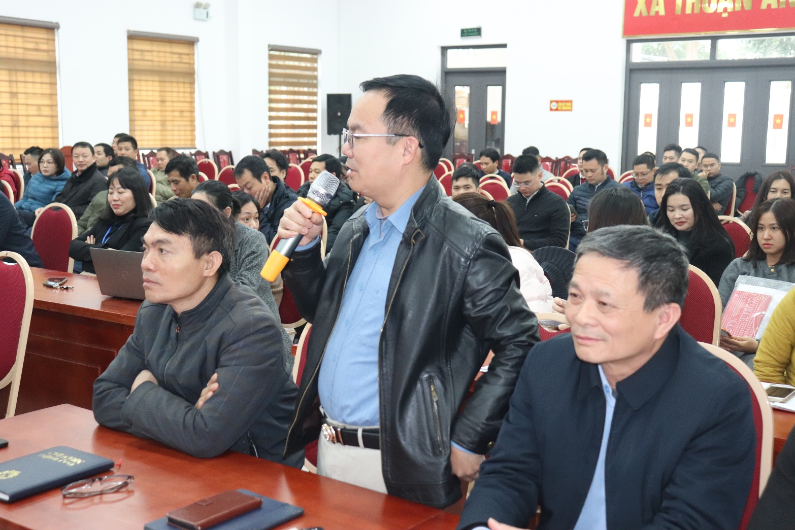 A person speaking into a microphone in front of a group of people  AI-generated content may be incorrect.