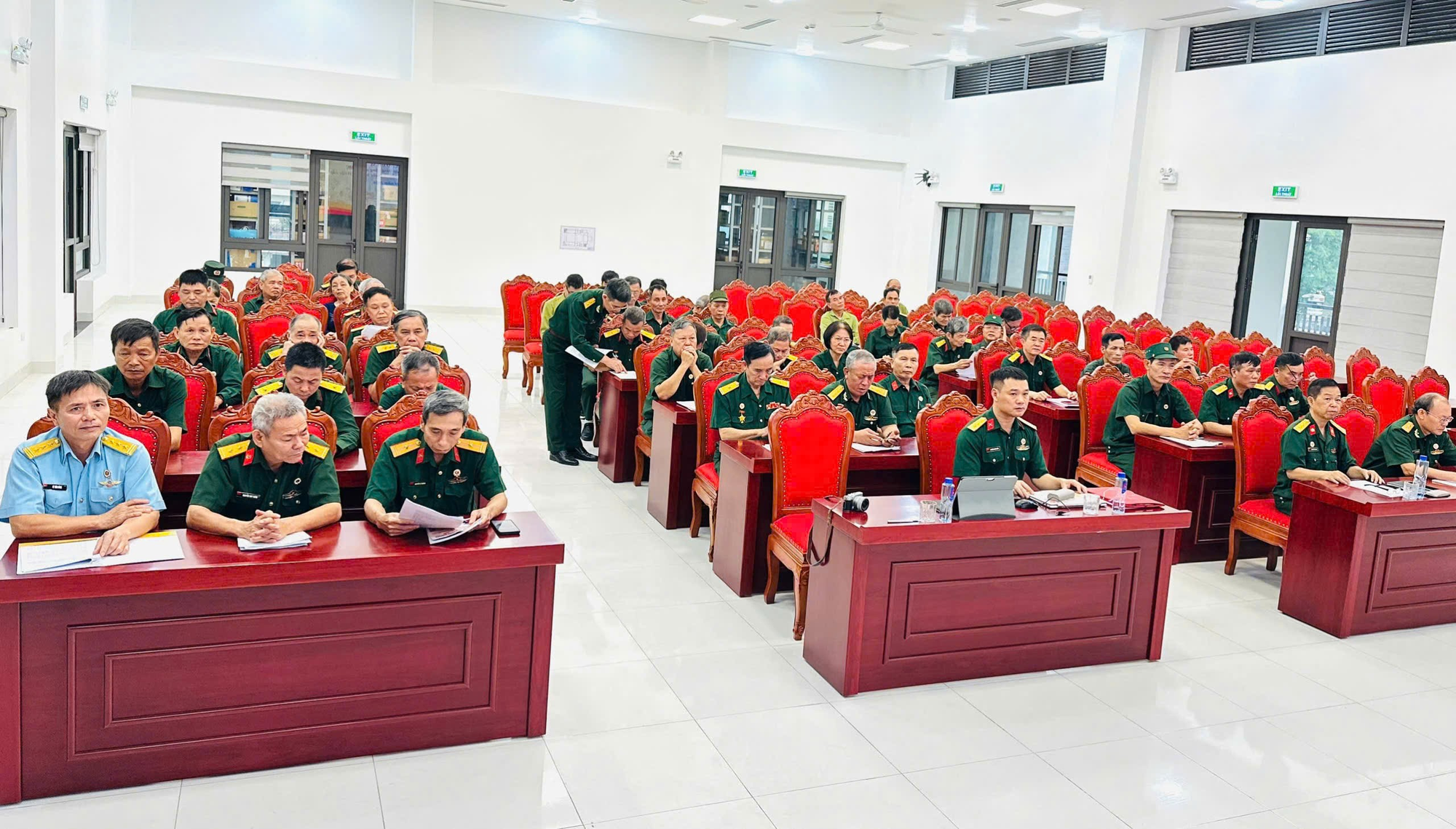 The image shows a room with a long table filled with chairs, where a group of people in uniformed green jackets are seated, and a large wooden desk is positioned at the front, surrounded by a neatly organized office environment.  AI-generated content may be incorrect.