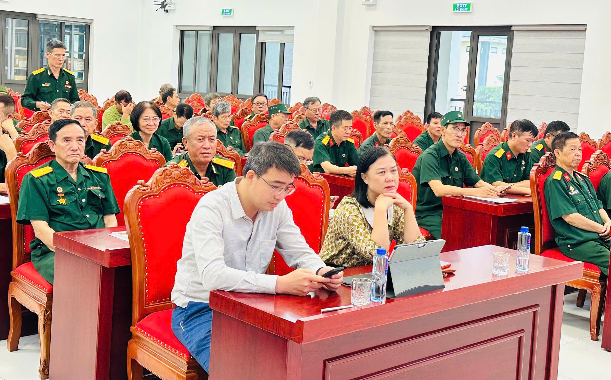 The image depicts a classroom setting with a large group of people, likely students or attendees, seated in rows of chairs. They are dressed in green uniforms and are engaged in using laptops or tablets, suggesting a classroom activity or presentation. The room has large windows, allowing natural light to fill the space, and the seating arrangement is formal, indicating an organized, structured environment.  AI-generated content may be incorrect.