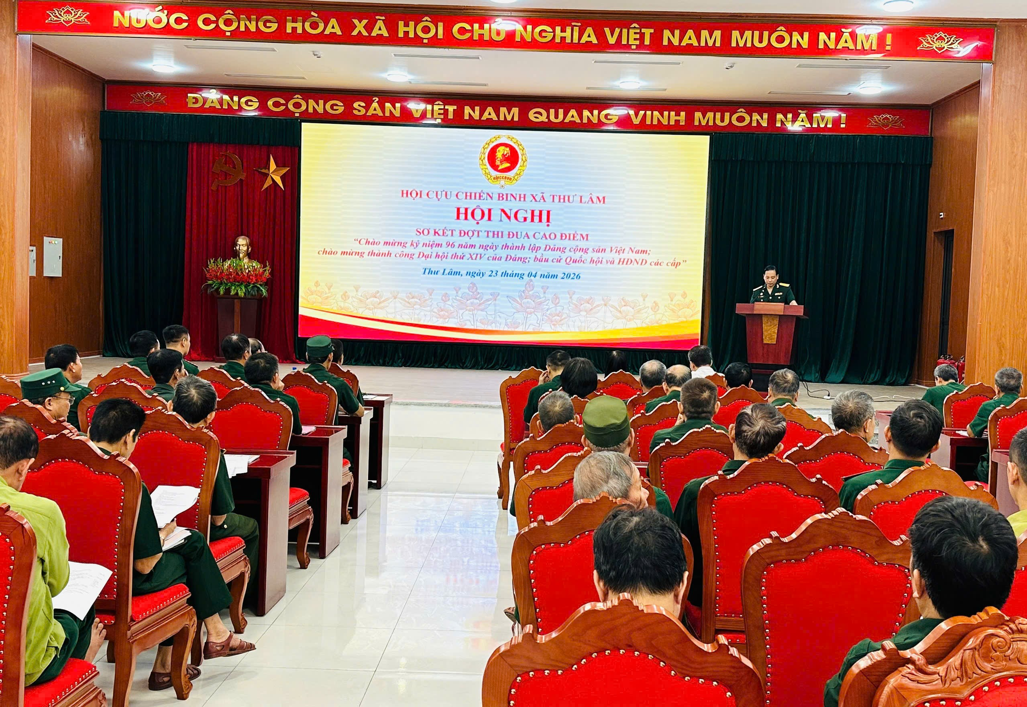 The image shows a large room with rows of chairs, a podium, and a decorated stage. A speaker stands at the podium, reading from a document in Vietnamese. The document is a commemorative speech, acknowledging the 96th anniversary of the establishment of the Vietnamese Front and the 14th Congress of the Workers' Party of Vietnam.  AI-generated content may be incorrect.