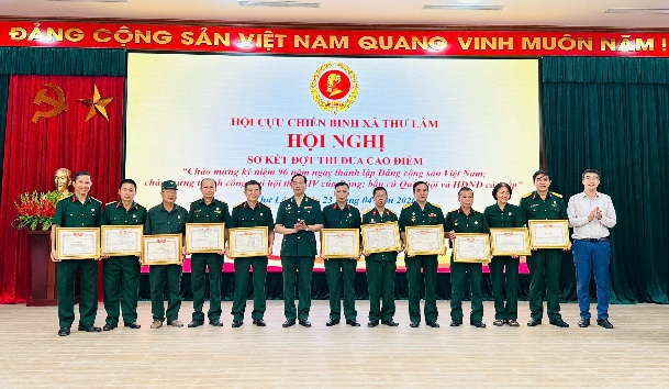 The image depicts a group of Vietnamese officials and dignitaries standing in a formal setting, dressed in military uniforms, and holding certificates or awards, with a red banner and the Vietnamese national flag in the background.  AI-generated content may be incorrect.